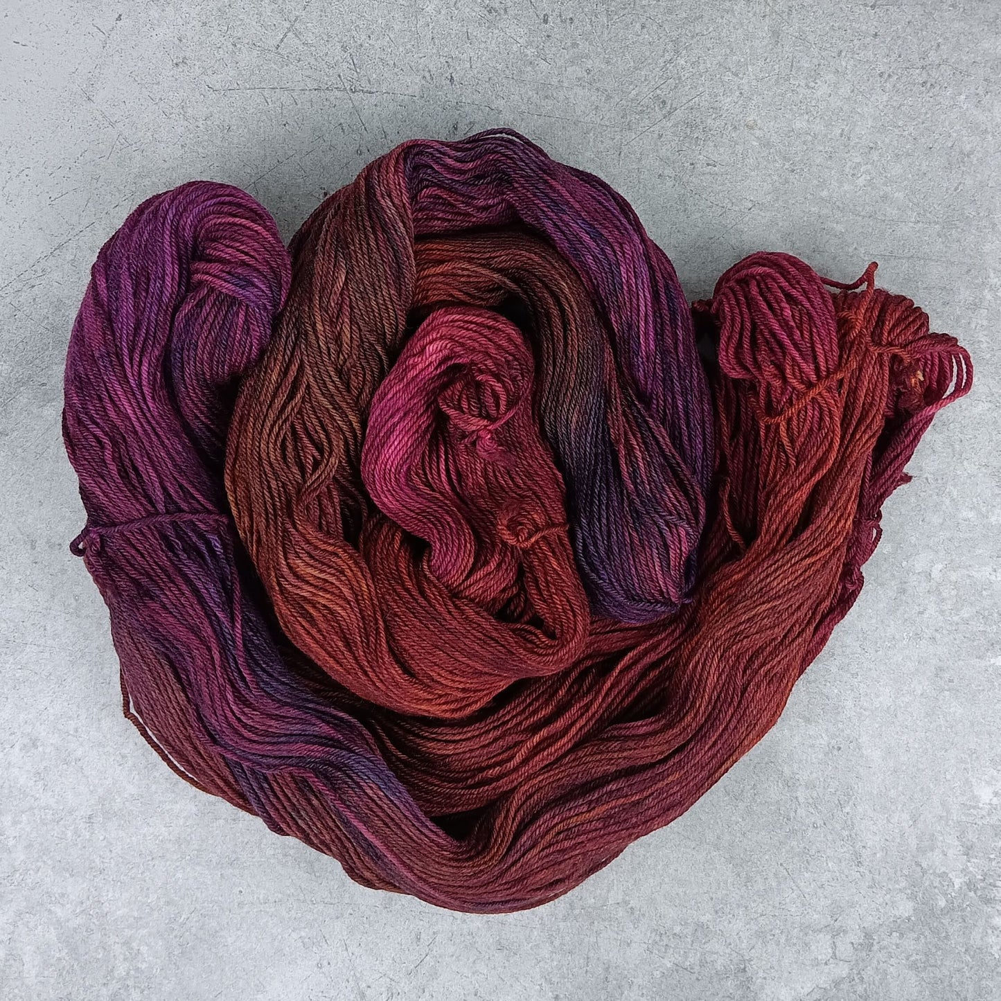 Two skeins of multicolored yarn in a maroon and purple variegated colourway on a gray surface