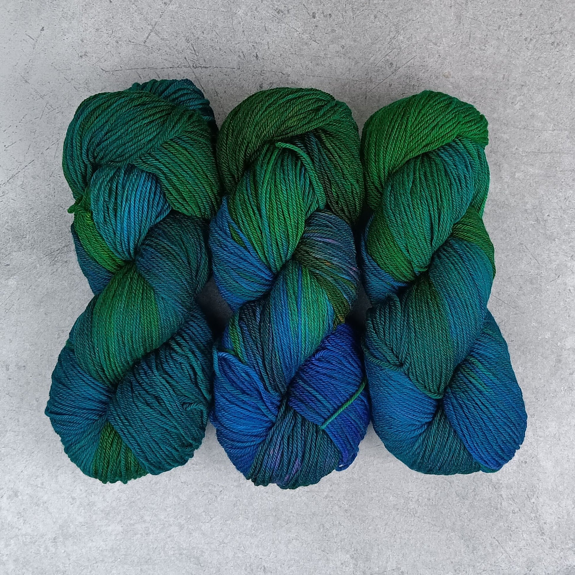 Three skeins of multicolored yarn in a variegated colourway with deep greens, blues, and little of purple on a gray surface