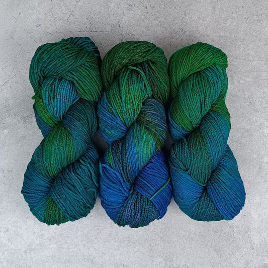 Three skeins of multicolored yarn in a variegated colourway with deep greens, blues, and little of purple on a gray surface