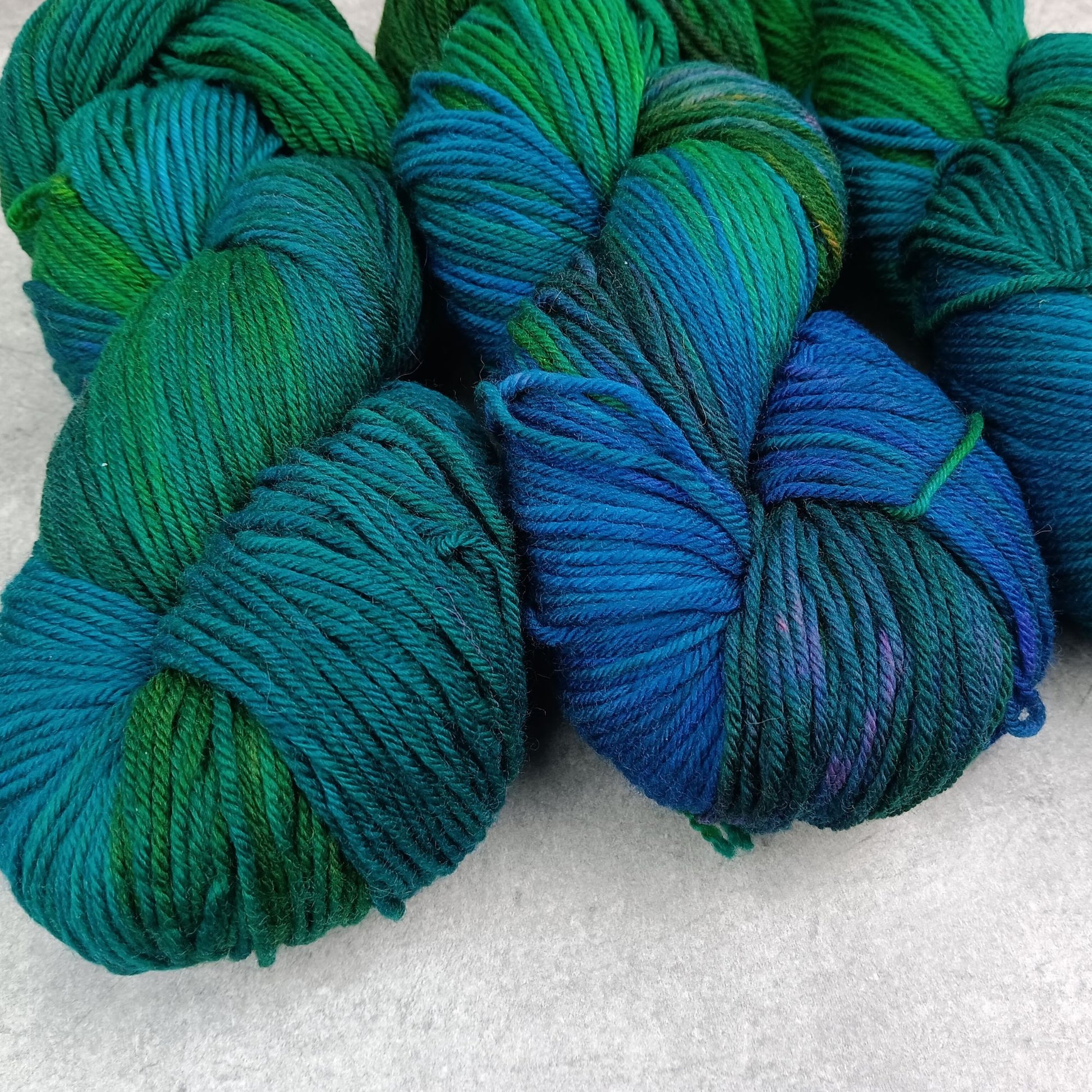 Three skeins of multicolored yarn in a variegated colourway with deep greens, blues, and little of purple on a gray surface