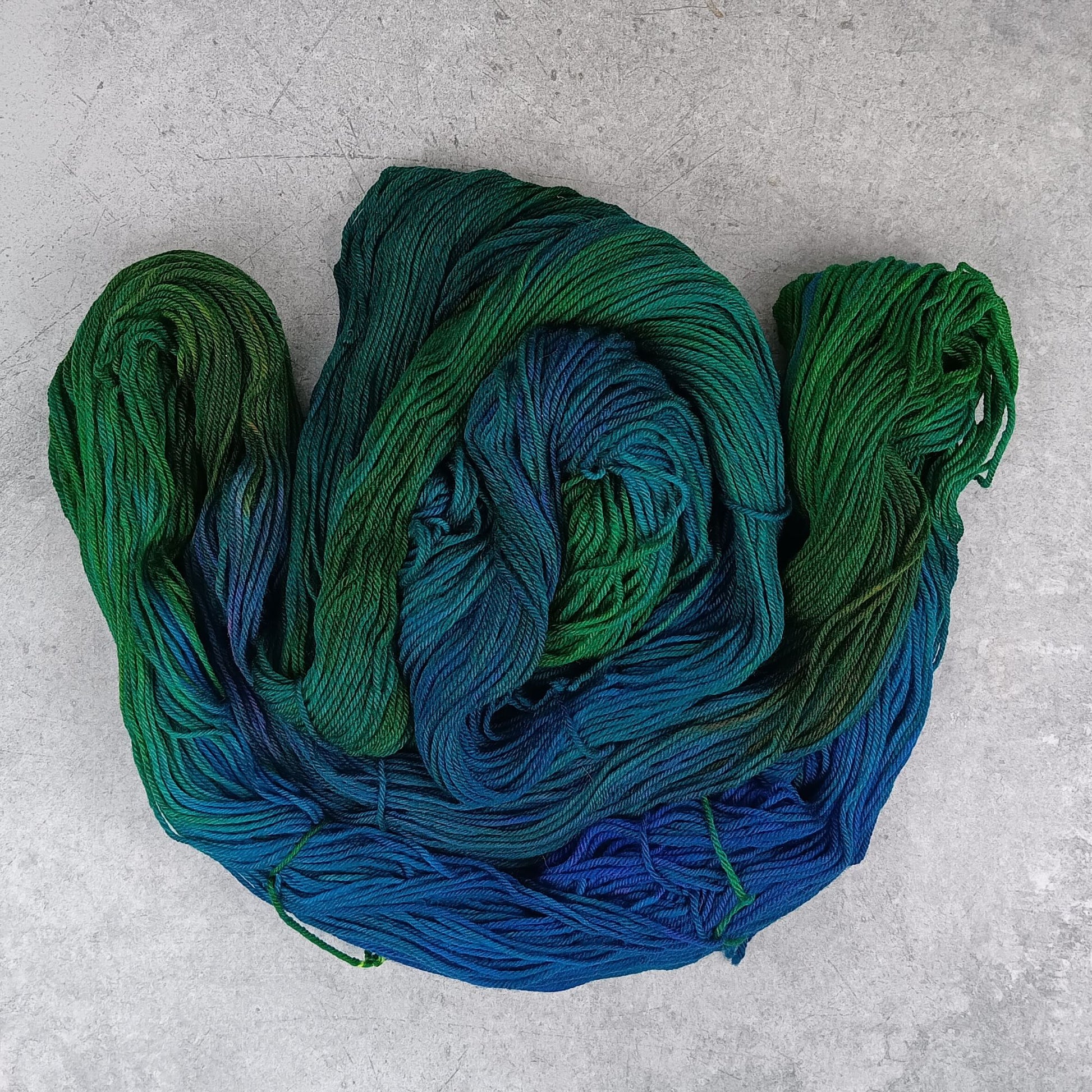 Three skeins of multicolored yarn in a variegated colourway with deep greens, blues, and little of purple on a gray surface